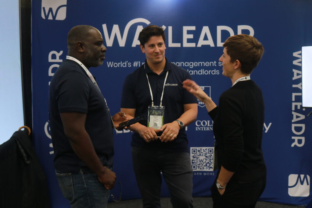 Three people stand and converse in front of a blue Wayleadr backdrop at a conference. Two are wearing navy blue shirts with lanyards, while the third person is in black.