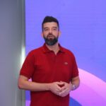 A man with a beard in a red shirt stands in front of a purple backdrop, speaking with his hands slightly raised.