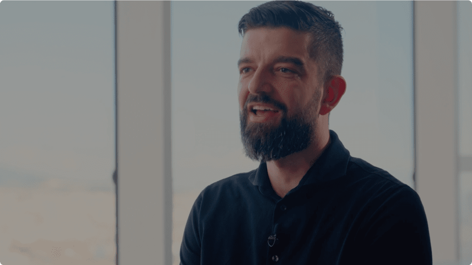 A man with a beard and short hair smiles while sitting indoors with large windows in the background.