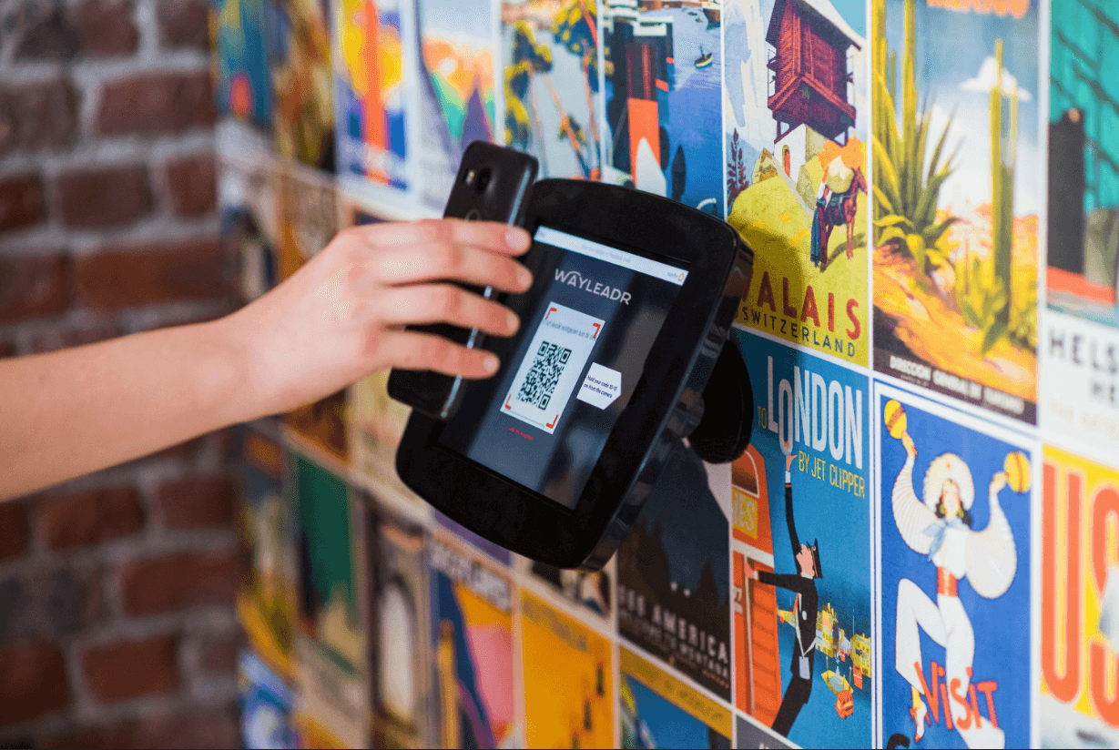 A person scans a QR code on a wall-mounted device displaying "Wayleadr" against a colorful poster-covered wall.