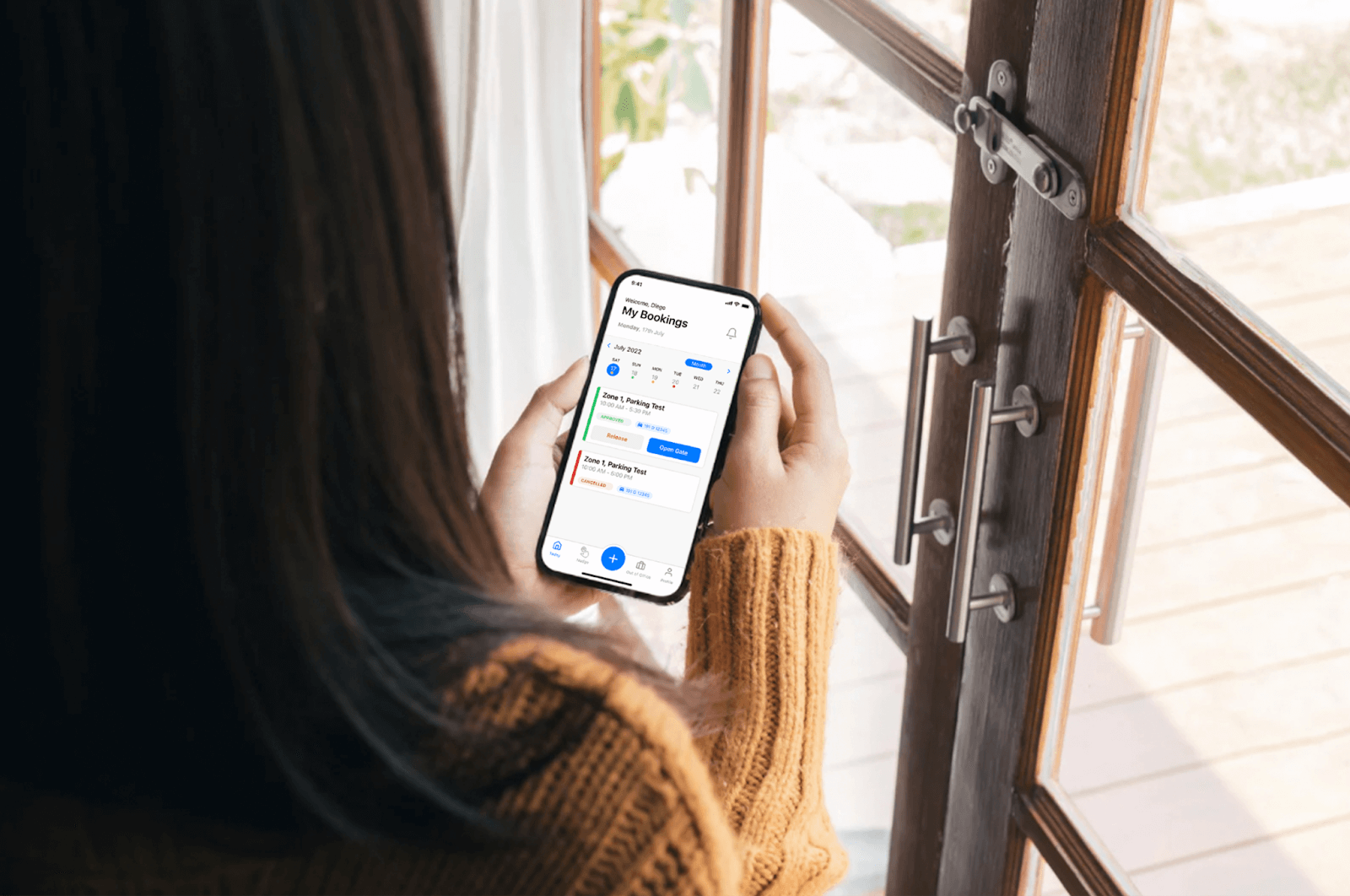 Person holding a smartphone with a calendar app open, standing by a wooden-framed window.