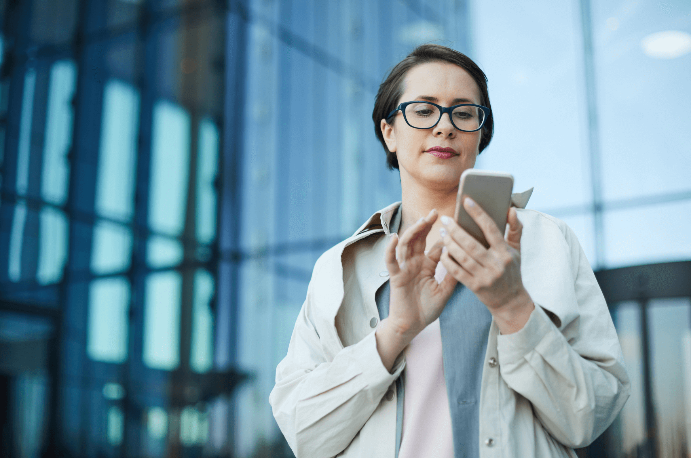 A person in a beige coat and glasses looks at their smartphone outside a glass building.