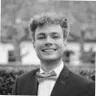 Black and white portrait of a person smiling, dressed in a suit and bow tie, with blurred trees in the background.