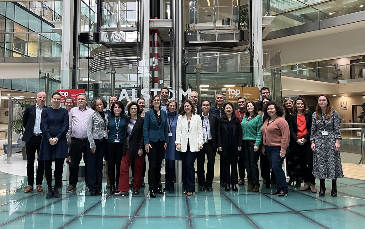 A group of people stands in front of an Alstom office building, posing for a group photo indoors.
