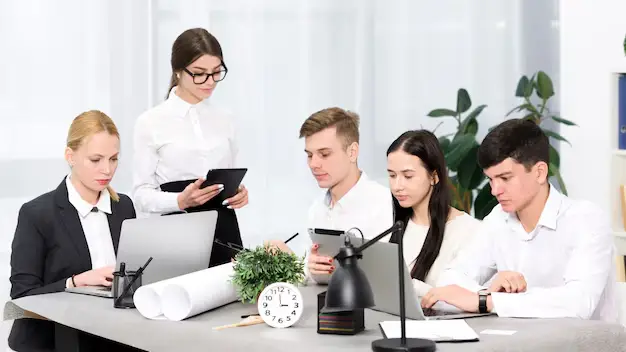 Five people in business attire sit and work at an office table with laptops, papers, plants, and a clock; one person stands holding a tablet.