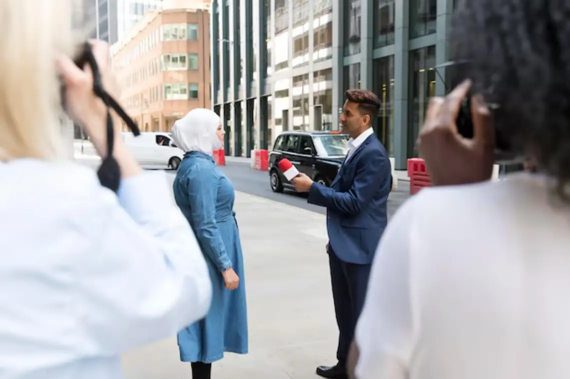 A man in a suit interviews a woman wearing a hijab and denim coat on a city street, while two people take photos of them.