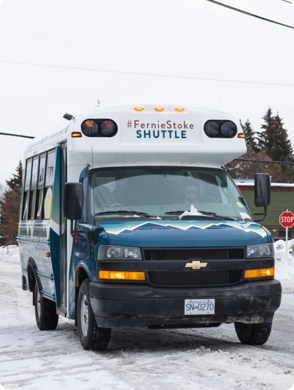 A blue and white shuttle bus labeled "#FernieStoke SHUTTLE" is parked on a snowy street with trees and houses in the background.
