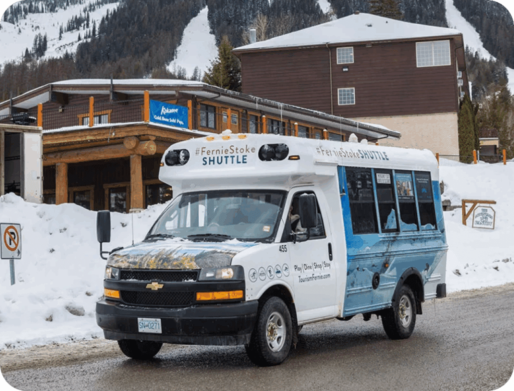 A FernieStoke Shuttle bus is parked on a snowy street in front of a wooden building with snow-covered mountains in the background.