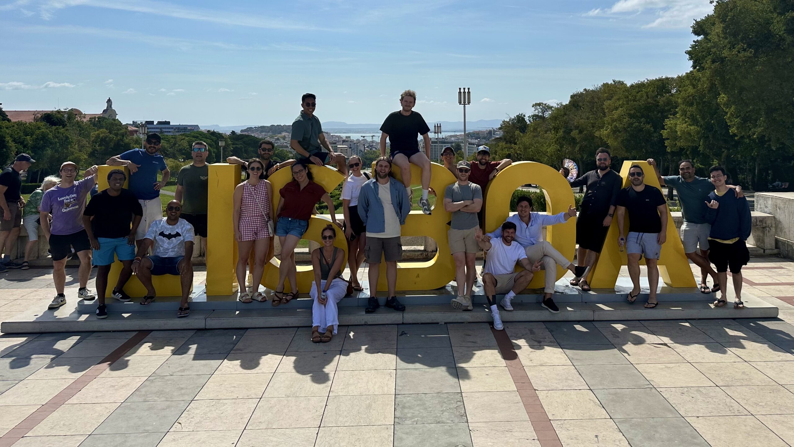 A group of people pose around and on top of large yellow "LISBOA" letters outdoors on a sunny day.