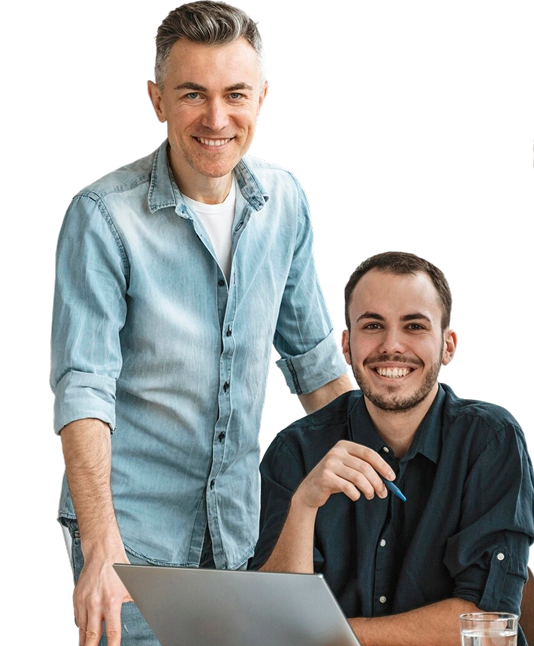 Two men smiling at the camera, one standing and one sitting at a desk with a laptop and a pen in hand.