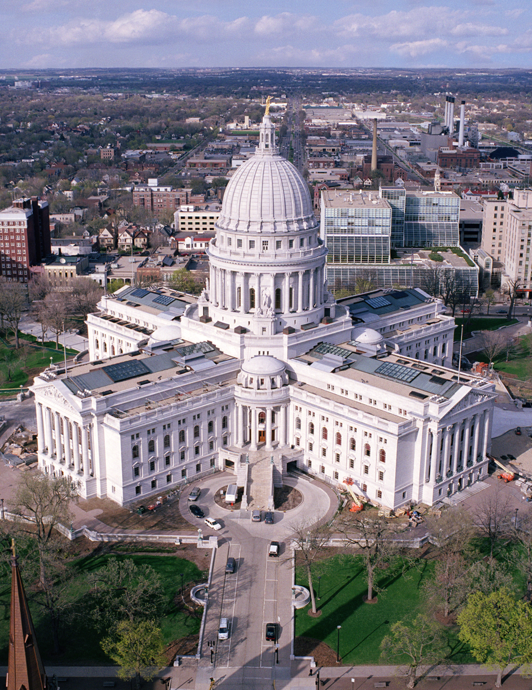 Aerial view of the Wisconsin State Capitol building with its white dome, surrounded by city streets and green trees, under a partly cloudy sky.