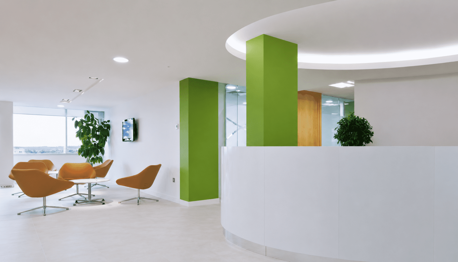 Modern office reception area with a curved white desk, green columns, orange chairs around a table, large windows, and potted plants.