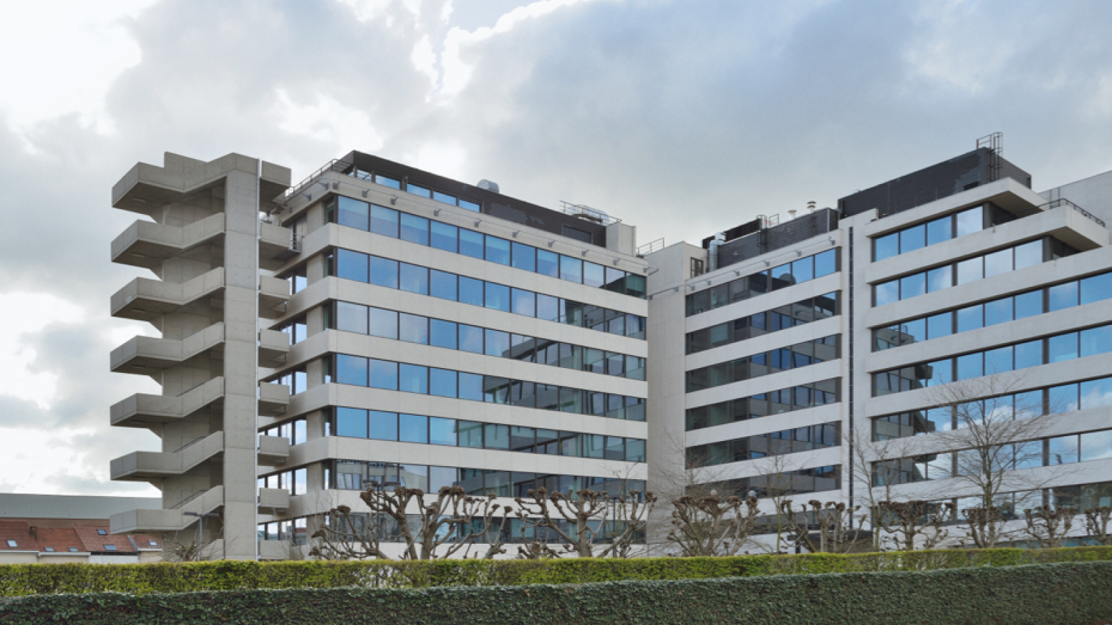 A modern office building with multiple floors, glass windows, and an external concrete staircase, set against a cloudy sky. Shrubs line the front of the building.