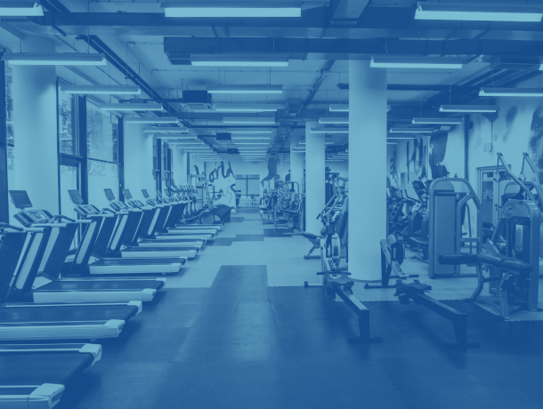 A spacious indoor gym with multiple treadmills, exercise bikes, and strength training equipment lined up neatly under bright fluorescent lighting.